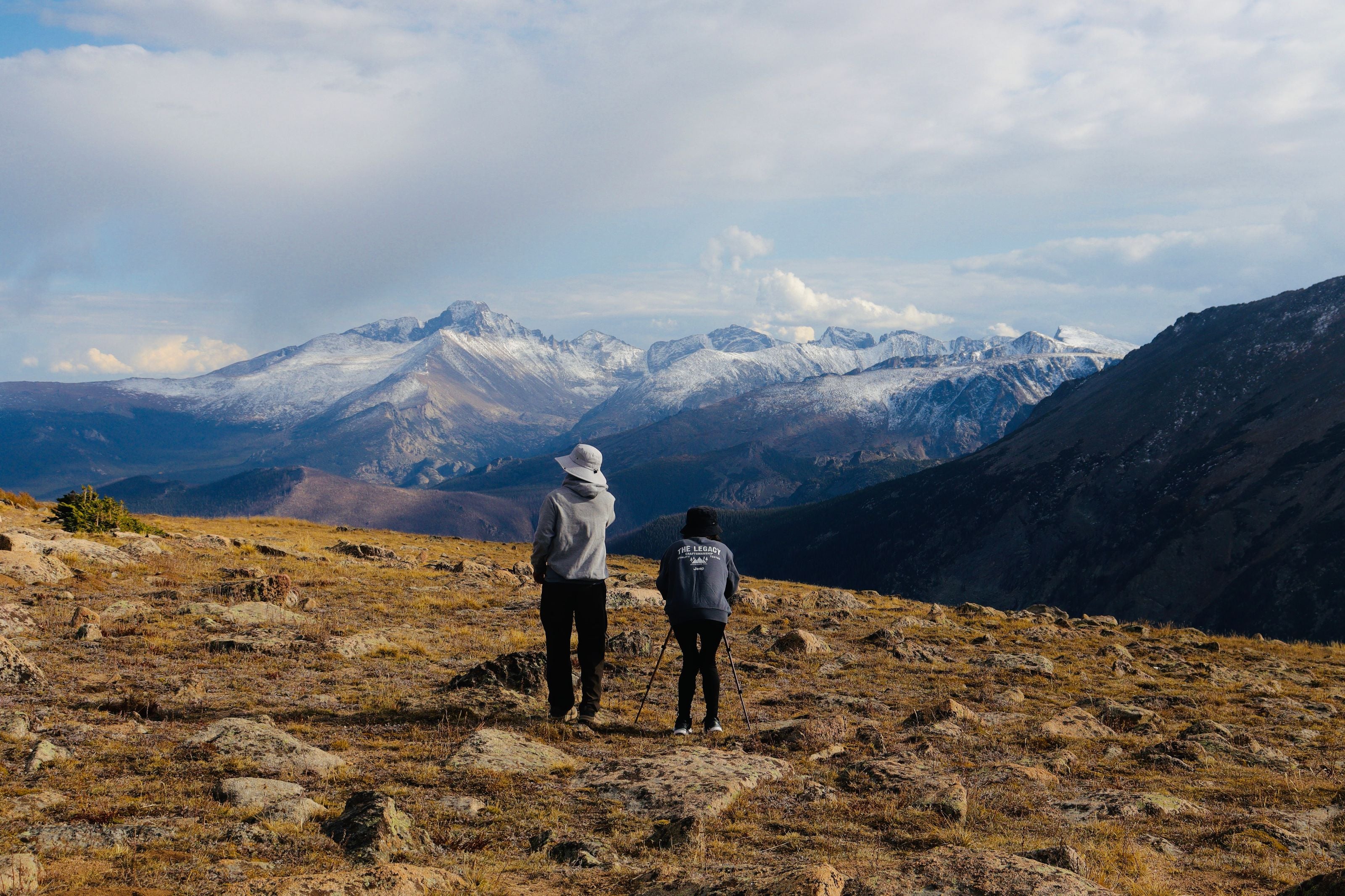 Two hikers in hoodies look at a snow-covered mountain range under a cloudy sky.