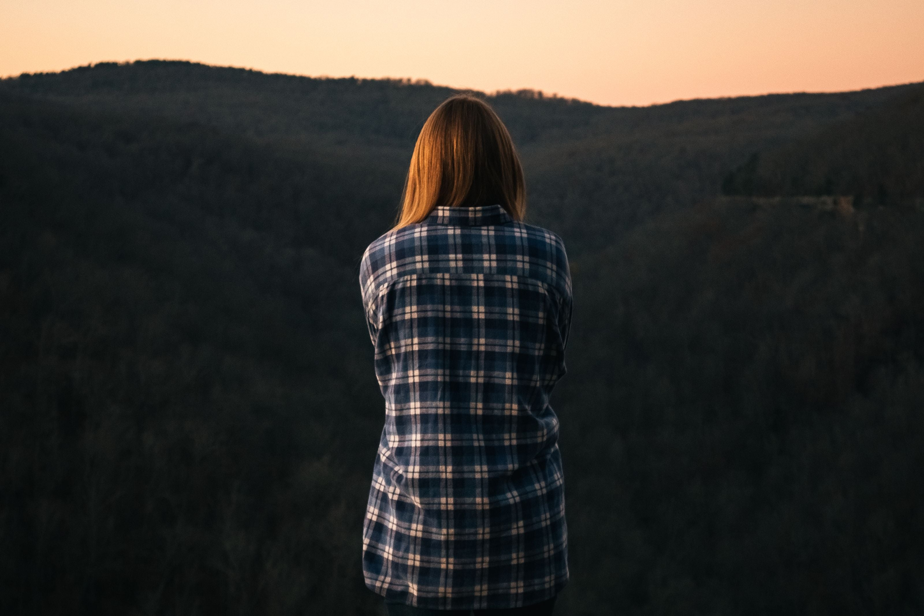 Woman in plaid shirt standing on a mountain during sunset, capturing solitude and nature's beauty.